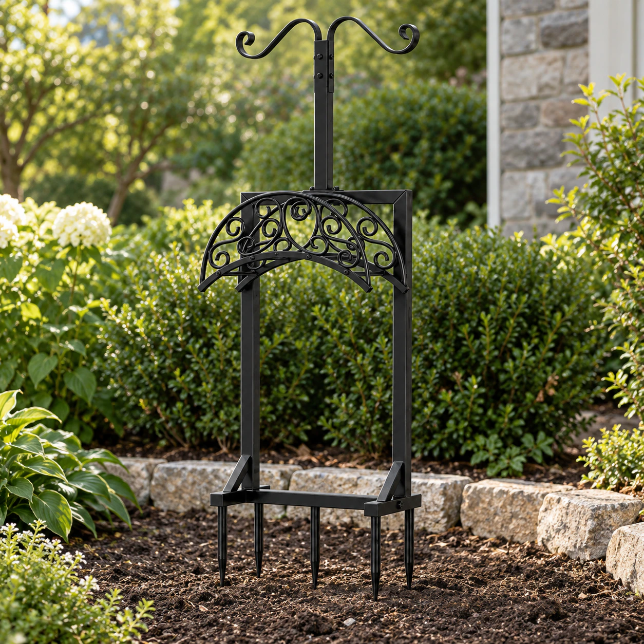 Black scrollwork hose stand with five visible ground prongs in a planted garden border